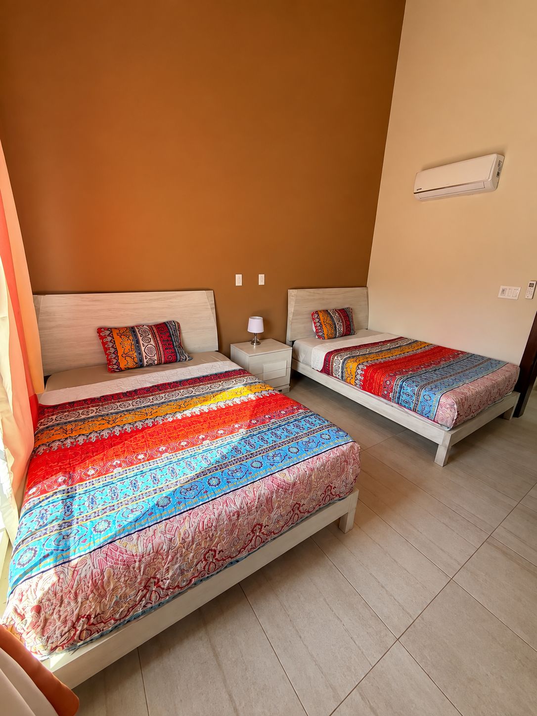 Quarto com duas camas, cada uma com roupa de cama estampada, mesa de cabeceira com abajur e um aparelho de ar condicionado na parede.