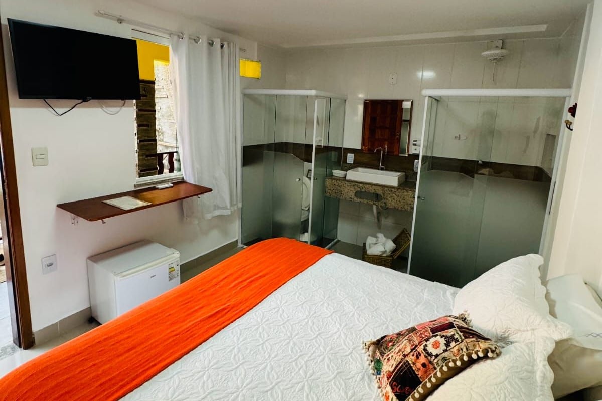 Ett hotellrum med en säng, en TV, ett litet kylskåp och ett badrum med en duschkabin med glasväggar.