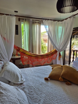 A bedroom with a bed, curtains, a hammock, and a view of greenery outside.