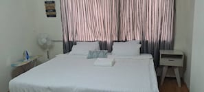 Una cama bien hecha con sábanas blancas, una mesita de noche con lámpara y un ventilador de pared.