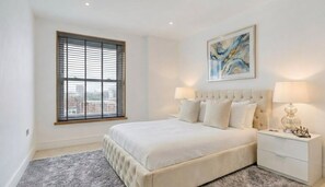 A bedroom with a large bed, two bedside tables, a window with blinds, and a painting on the wall. - Premium 1-Bedroom Apartment Greenwich Near O2 & Transport (London)