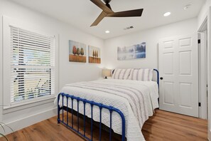 Quarto com cama, ventilador de teto, janela com persianas e obras de arte na parede.