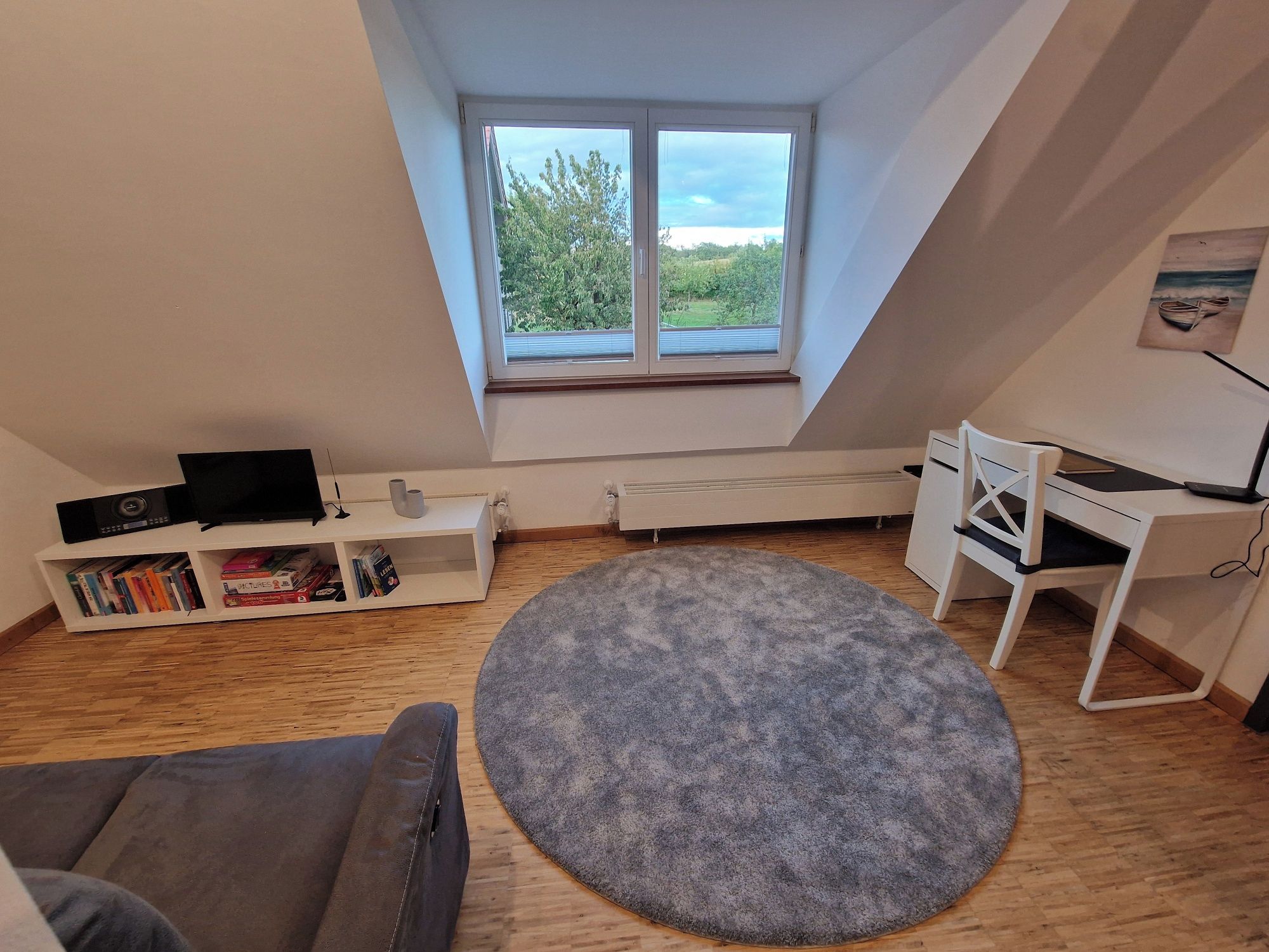 Ein Zimmer mit schrägem Dach, einem großen Fenster, einem weißen Schreibtisch, einem grauen Sofa und einem runden Teppich.