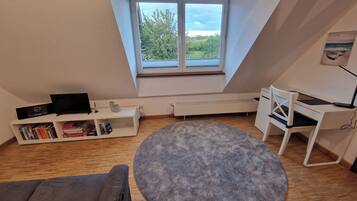 Ein Zimmer mit schrägem Dach, einem großen Fenster, einem weißen Schreibtisch, einem grauen Sofa und einem runden Teppich.