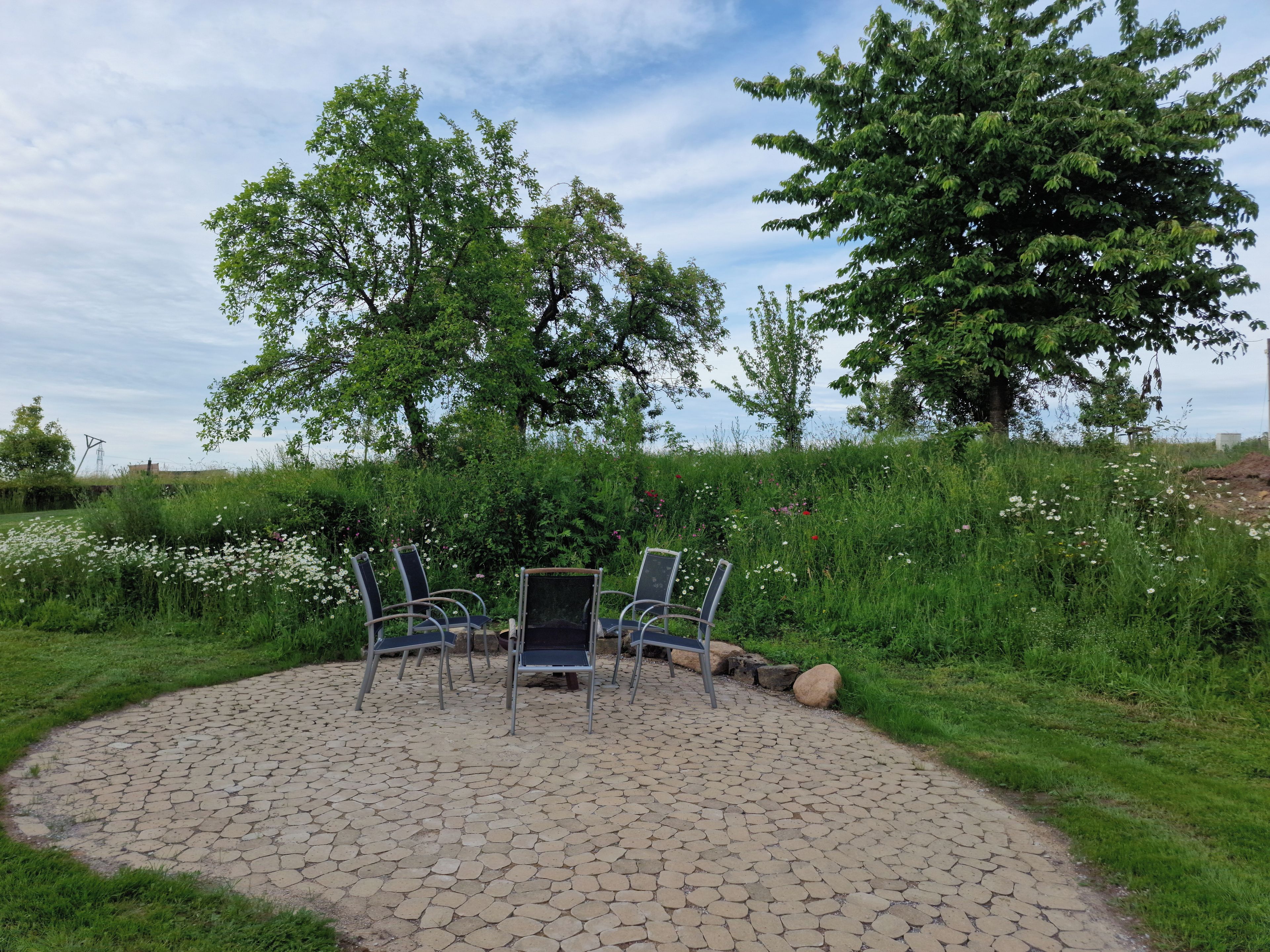 Un espace extérieur aménagé avec quatre chaises et une table, entouré d’un cercle pavé de pierres, avec des arbres et un champ d’herbe en arrière-plan.