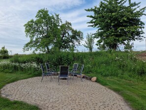 Ein Außenbereich mit Sitzgelegenheiten, vier Stühlen und einem Tisch, umgeben von einem kreisförmigen, mit Steinplatten befestigten Bereich, mit Bäumen und einem Rasen im Hintergrund.