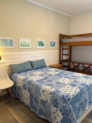 Ein Schlafzimmer mit Etagenbett, einem Bett mit blumigem, blauem Überwurf, einem weißen Sessel und vier gerahmten Pflanzenbildern an der Wand.