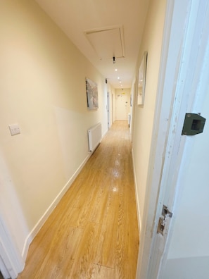 A hallway with wooden flooring, white walls, and a door with a black handle. - Heathrow Tranquil 4-bedroom apartment in delightful Feltham perfect for families (Feltham)