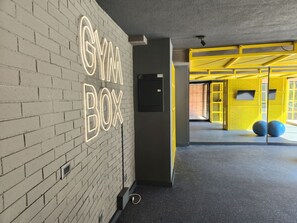 Ein Fitnessstudio mit einer Neonreklame mit der Aufschrift 'GYM BOX' an einer Backsteinwand, einer Tür mit Schloss und einem gelben Spielbereich mit Trainingsbällen.