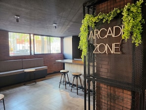 Un intérieur moderne avec un espace bar appelé « ZONE ARCADE », comprenant un comptoir en bois, des tabourets et un coin salon avec un banc et des tables.