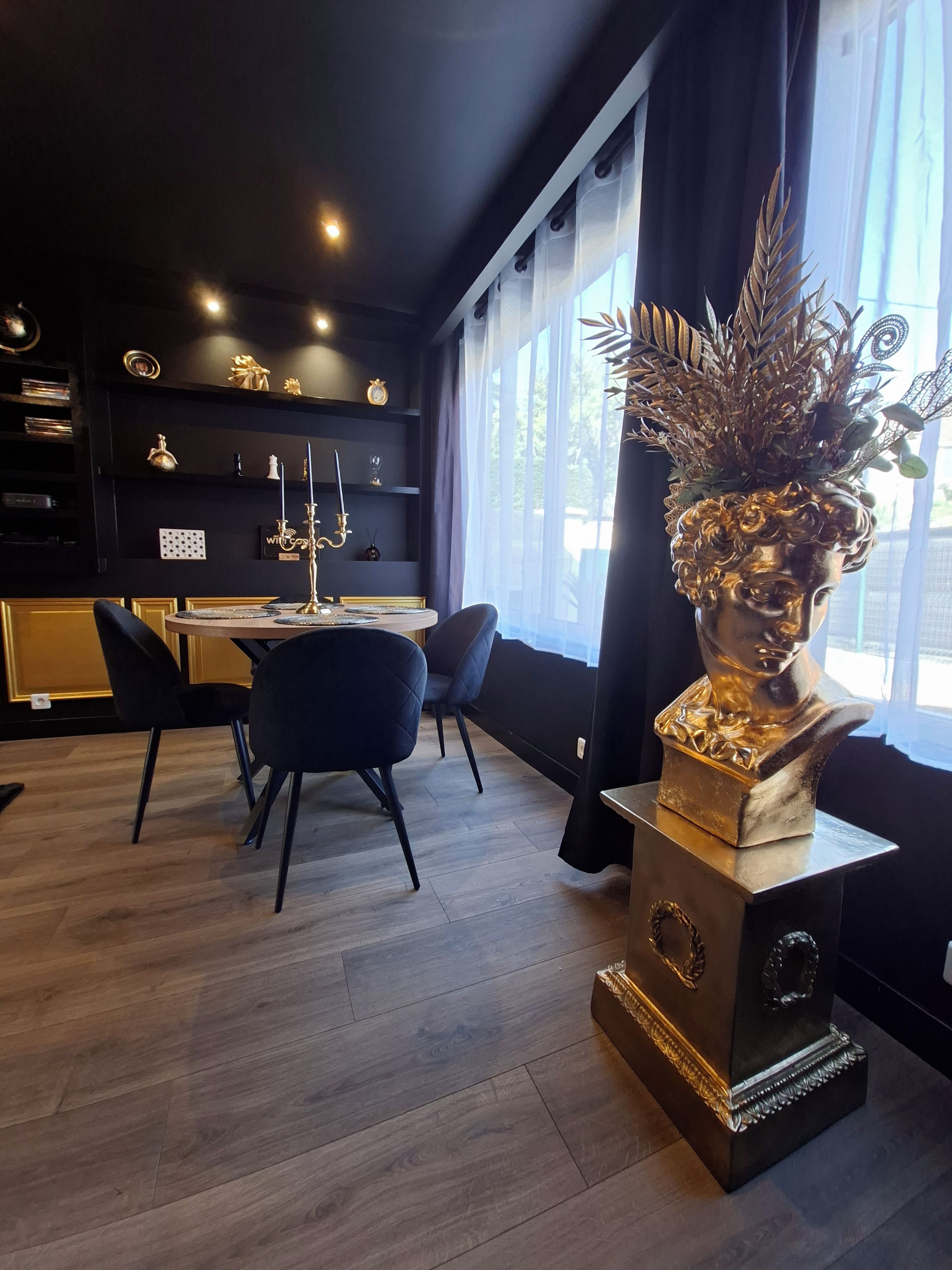 Un espace de salle à manger moderne aux couleurs noir et or, agrémenté d’un lustre, d’un vase orné d’un buste doré et offrant une vue sur l’extérieur grâce à de grandes fenêtres.