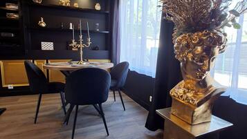 Un espace de salle à manger moderne aux couleurs noir et or, agrémenté d’un lustre, d’un vase orné d’un buste doré et offrant une vue sur l’extérieur grâce à de grandes fenêtres.