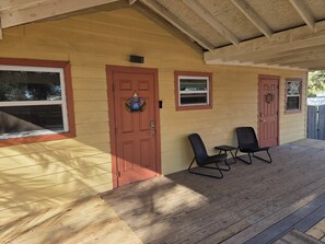Un porche couvert avec un plancher en bois, deux chaises noires et une porte rouge ornée d’une pancarte indiquant « Bienvenue ».