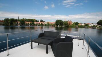 Eine Terrasse mit einer Couch und einem Tisch, mit Blick auf einen ruhigen Fluss und Gebäude im Hintergrund.
