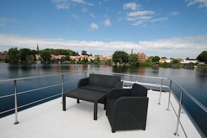 Una terraza con sofá y mesa, con vistas a un río tranquilo y edificios al fondo.