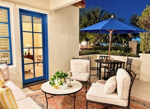 An outdoor patio with a dining table, chairs, a blue umbrella, and a view of the garden. - A Charming Legacy Villa
3 bedroom 4 bath Steps to Pool & Stunning Mountain Views (La Quinta)