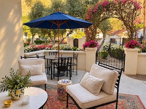 An outdoor patio with a round table, two chairs, a blue umbrella, and a potted plant. - A Charming Legacy Villa
3 bedroom 4 bath Steps to Pool & Stunning Mountain Views (La Quinta)