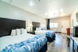 A hotel room with two beds, a desk, a refrigerator, and a window with curtains. - ATRIA INN & SUITES EXTENDED STAY  (Jourdanton)