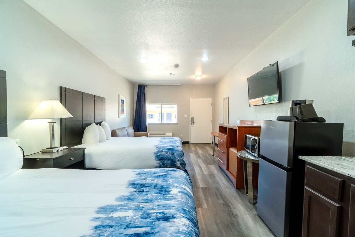 A hotel room with two beds, a desk with a lamp, a microwave, a refrigerator, and a television on the wall.