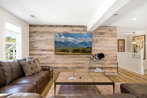 A living room with a wooden wall, a mounted picture of mountains, a leather sofa, a coffee table, and a staircase with a railing. - Walk to Ski Trolley | Private Hot Tub | Sleeps 23 (Park City)