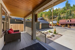 A covered porch with a wicker chair and a red cushion, a wooden fence, and a view of a street with buildings and trees. - Walk to Ski Trolley | Private Hot Tub | Sleeps 23 (Park City)