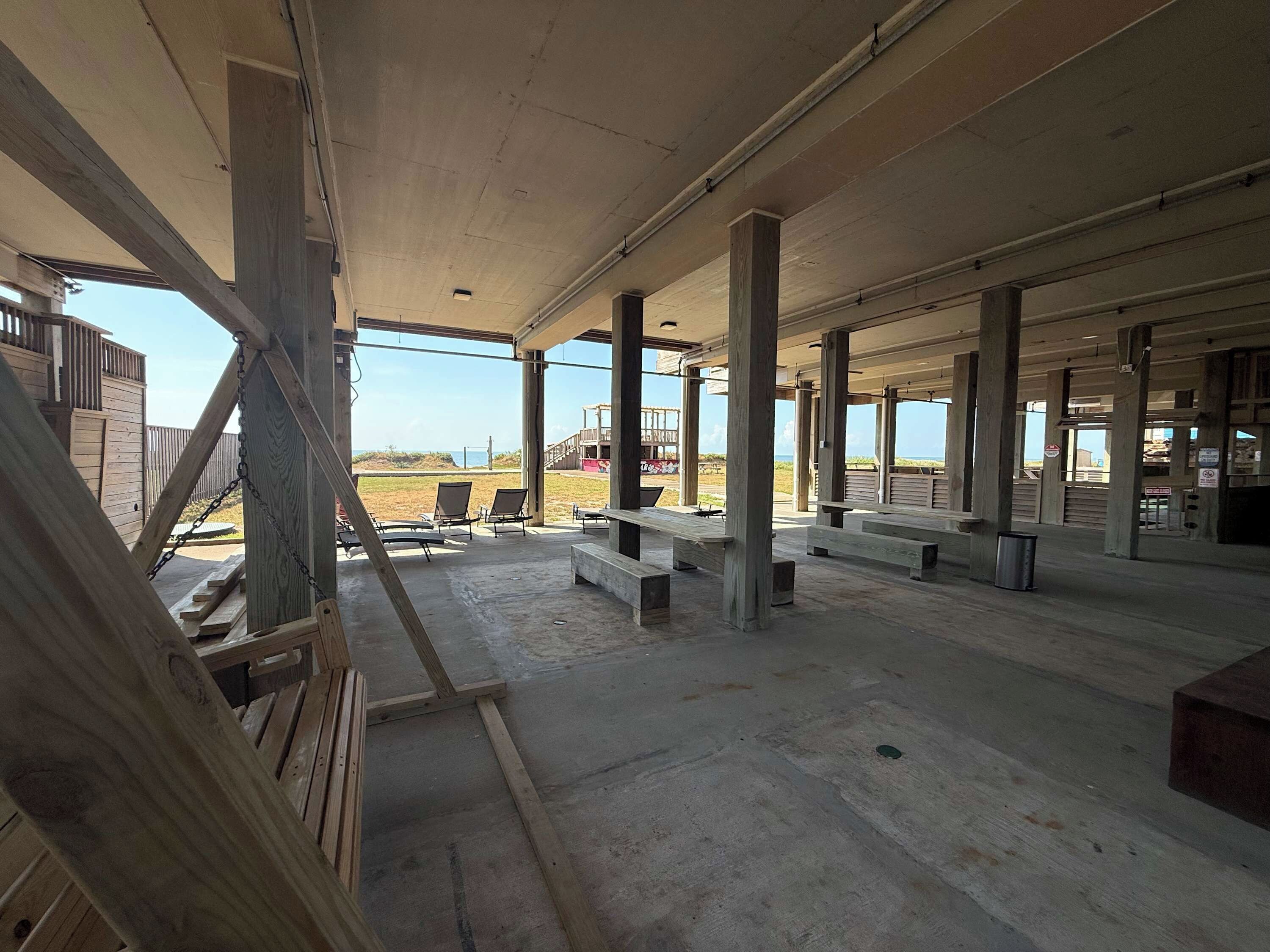 An open-air covered patio area with wooden benches and tables, concrete flooring, and a view of the beach.