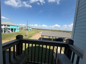 A view from a porch with a wooden railing, overlooking a grassy area with a building in the distance. - Casa Dora Pearl Beachfront (Bolivar Peninsula)