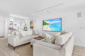 A modern living room with a sectional sofa, a coffee table with decorative items, a flat-screen TV mounted on the wall, and a kitchen area with built-in shelves. - Naples Bay Waterfront Dolphins, Boat Dock & Pool (Naples)