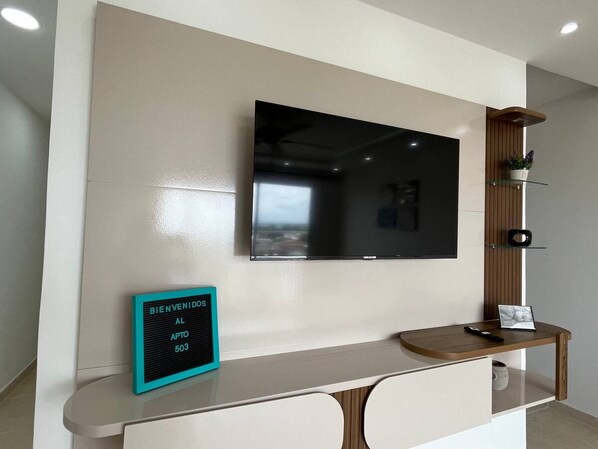 Un espace d’accueil doté d’un téléviseur à écran plat fixé au mur, d’une pancarte de bienvenue, d’une console en bois et d’une petite étagère ornée d’objets décoratifs.