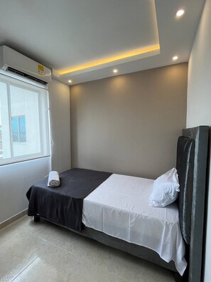 Un lit simple avec une couvre-lit noir et blanc, un oreiller blanc et une tête de lit noire, dans une pièce dotée d’une fenêtre et d’un éclairage encastré au plafond.