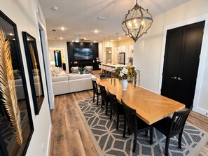 A modern living room with a sofa, a dining table with chairs, a TV, and a kitchen area in the background. - Shell We Vacay 30A-Spacious 4 Bedroom - Private Saltwater Pool, PCB Florida (Panama City Beach)