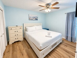 A bedroom with a bed, nightstand, dresser, ceiling fan, and artwork on the wall. - Coastal 2bd Condo with Community Pool and Hot Tub (Panama City Beach)