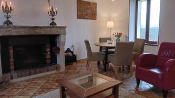 A living room with a fireplace, a dining table with chairs, a red armchair, and a view through a window.