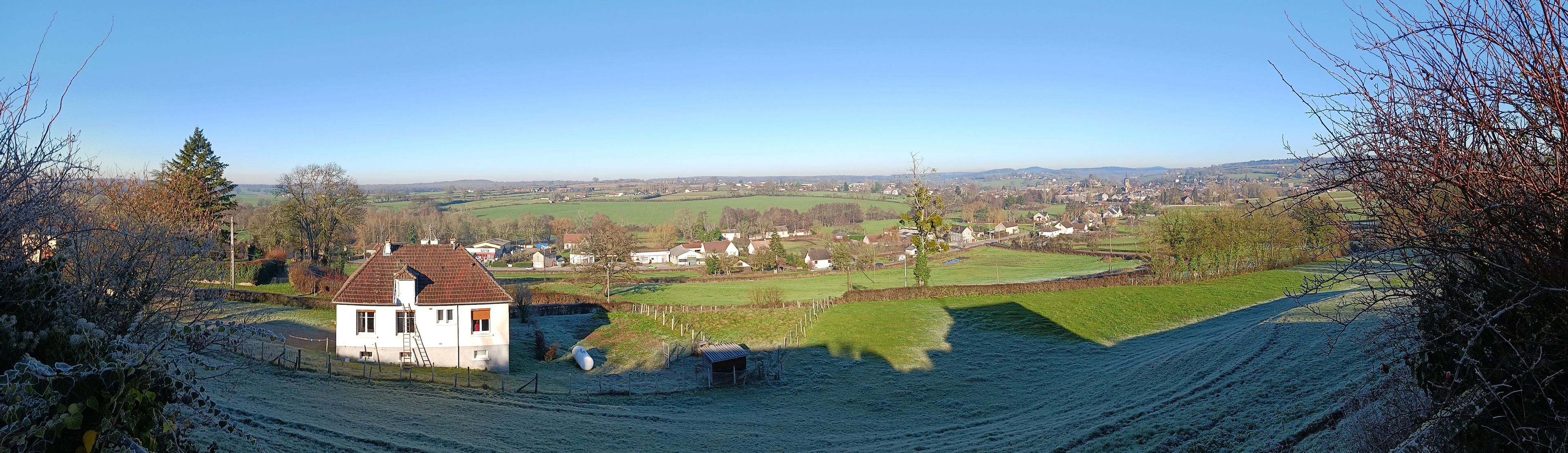 Un paysage rural avec une maison blanche, des champs verdoyants et une ville qui se devine au loin.