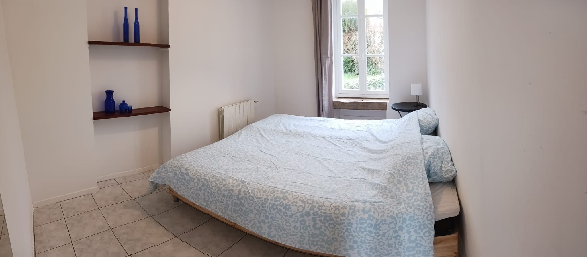 Ein Schlafzimmer mit einem Bett, einem Heizkörper, einem Fenster mit Vorhängen und einem kleinen Tisch mit einer Lampe.