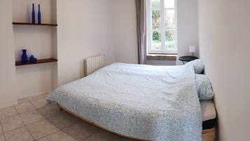 A bedroom with a bed, a radiator, a window with curtains, and a small table with a lamp.