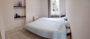 Ein Schlafzimmer mit einem Bett, einem Heizkörper, einem Fenster mit Vorhängen und einem kleinen Tisch mit einer Lampe.