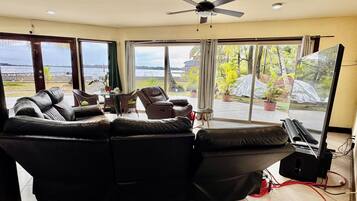 A living room with a black leather sofa, a ceiling fan, and a large window offering a view of the outdoors.