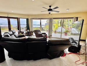 A living room with a black leather sofa, a ceiling fan, and a large window offering a view of the outdoors.