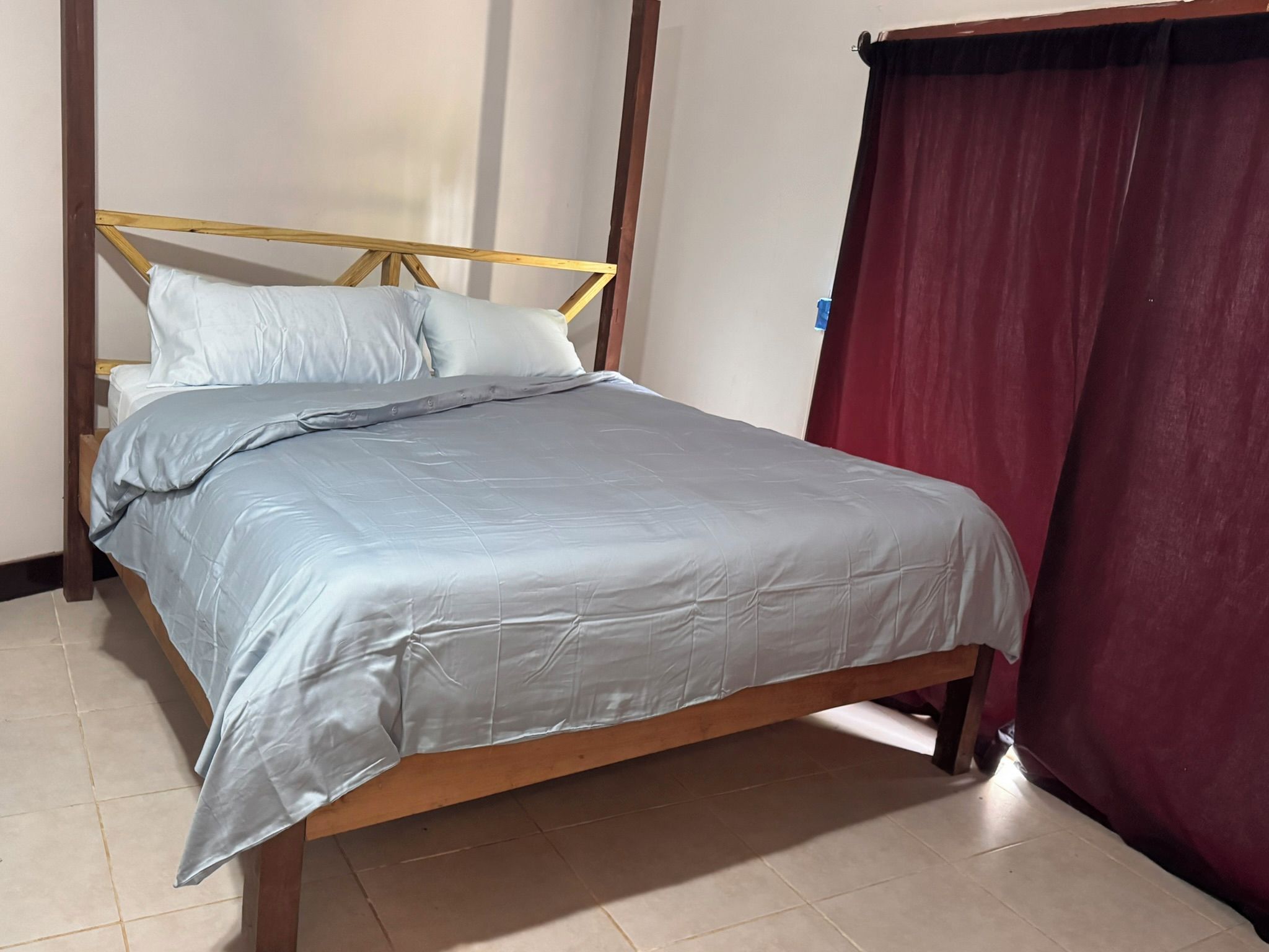 Cama de solteiro com estrutura de madeira e colcha cinza, encostada em uma parede com cortina vermelha.
