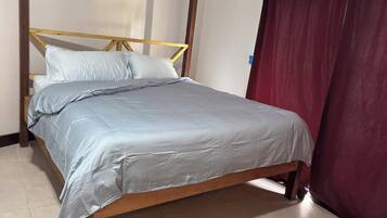 Cama de solteiro com estrutura de madeira e colcha cinza, encostada em uma parede com cortina vermelha.