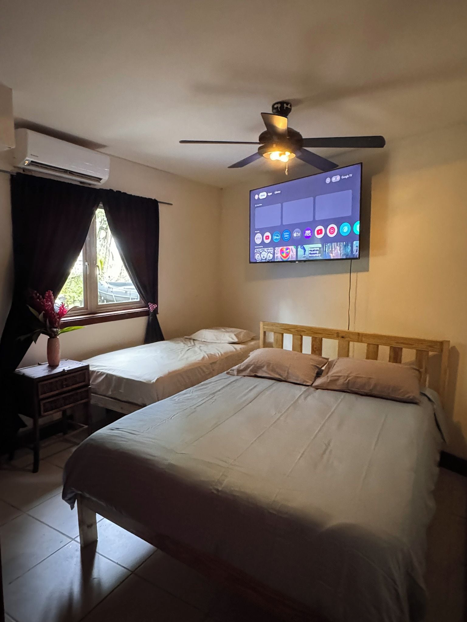 Quarto de hotel com duas camas, ventoinha no teto, ar condicionado, televisão presa à parede e janela com cortinas.