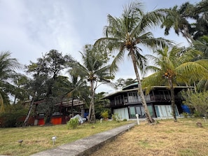 Un edificio de dos pisos con un balcón, rodeado de palmeras y un área verde.