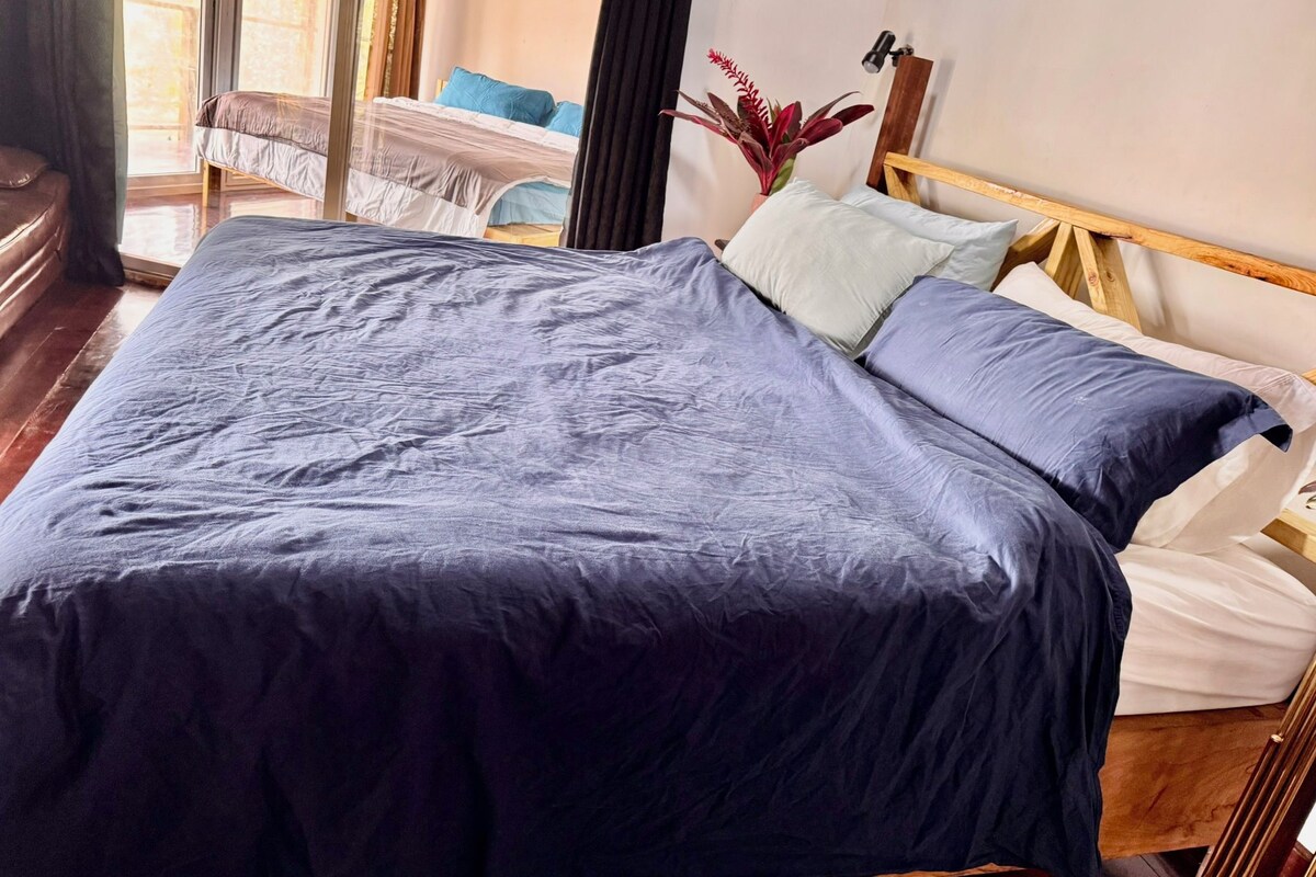 A bed with a dark blue bedspread and pillows, a wooden headboard, and a potted plant on the nightstand.