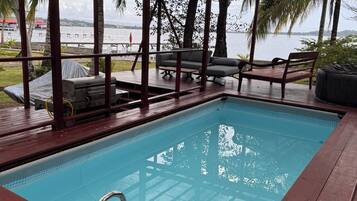 Una piscina con cubierta de madera y barandillas metálicas, rodeada de palmeras y vista al mar.
