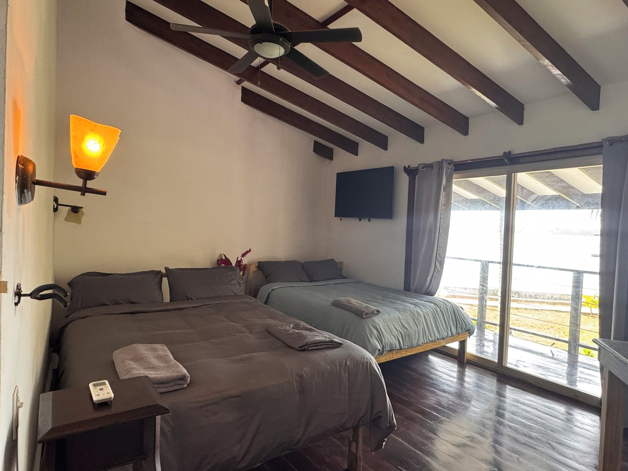 Una habitación con dos camas, ventilador de techo, televisor montado y ventana con cortinas.