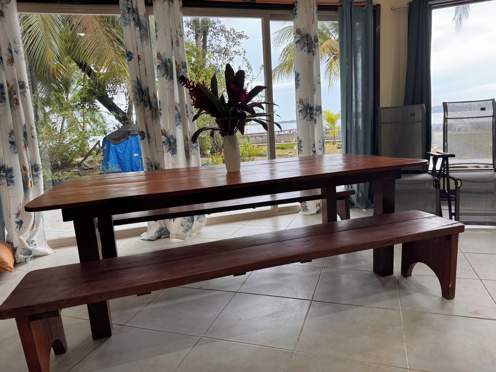 A wooden table with benches in a room with large windows and a view of palm trees.