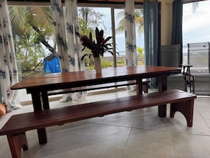 A wooden table with benches in a room with large windows and a view of palm trees.