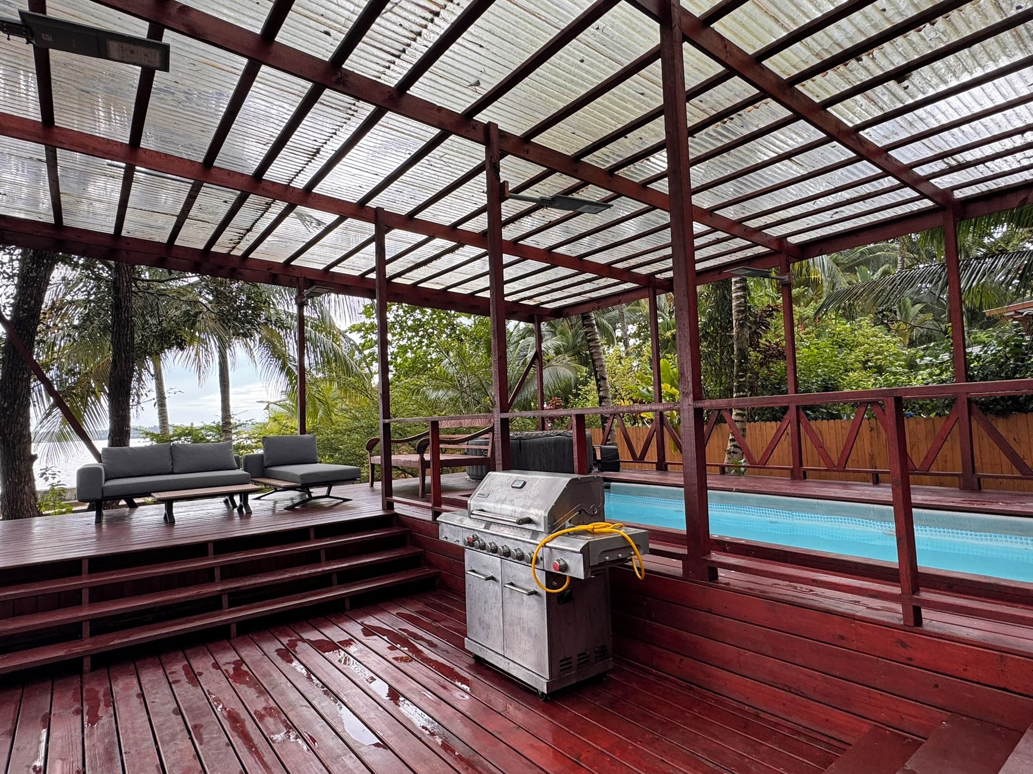 Un espacio al aire libre techado con piscina, terraza de madera, parrilla y asientos.