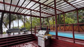 Un espacio al aire libre techado con piscina, terraza de madera, parrilla y asientos.